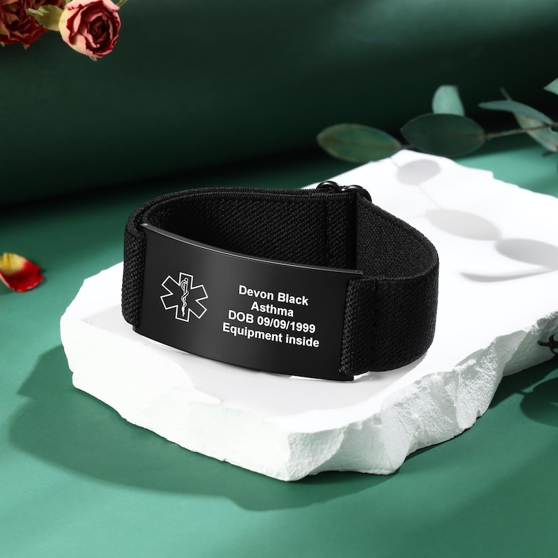 May include: Black medical alert bracelet with a black plate. The plate has a medical symbol and the following text: Devon Black, Asthma, DOB 09/09/1999, Equipment inside. The bracelet is on a white surface.