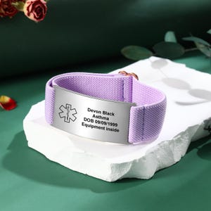 May include: A lavender medical ID bracelet with a silver plate. The plate has the Star of Life symbol and text that reads "Devon Black, Asthma, DOB 09/09/1999, Equipment inside." The band is made of a woven fabric.