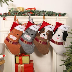 Peut inclure: Quatre bas de Noël en forme de chat et de chiens, chacun avec un bonnet de Père Noël. Les bas sont marron, gris et blancs, avec des ceintures rouges et les noms "Maya", "Mason", "Jack" et "Angel" brodés dessus.