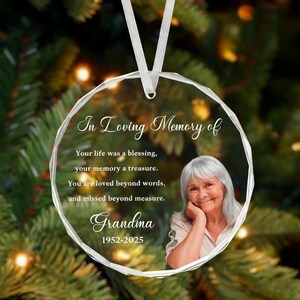 Puede incluir: Un adorno circular de cristal transparente con una cinta blanca. El texto dice In Loving Memory of, Your life was a blessing, your memory a treasure. You are loved beyond words, and missed beyond measure. Grandma 1952-2025, con la foto de una mujer.