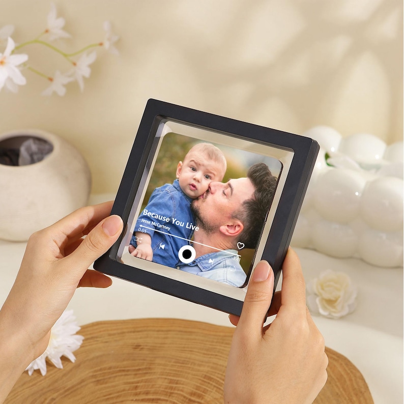 May include: A black square photo frame displaying a picture of a man kissing a baby. The image has the text "Because You Live" and a music player interface. The frame is held by two hands.