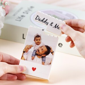 Puede incluir: Un marco de fotos blanco en forma de bloque con una foto de un padre y un ni&ntilde;o. El bloque superior dice "Daddy & Me" en escritura negra. Un coraz&oacute;n rojo est&aacute; impreso en la parte inferior de la foto. El marco lo sostiene una persona.