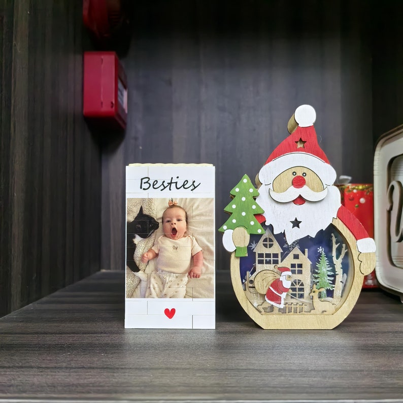 Puede incluir: Un bloque de fotos de madera con la palabra "Besties" y una foto de un beb&eacute;. Tambi&eacute;n hay una decoraci&oacute;n de madera de Pap&aacute; Noel con un &aacute;rbol de Navidad y una escena de pueblo.