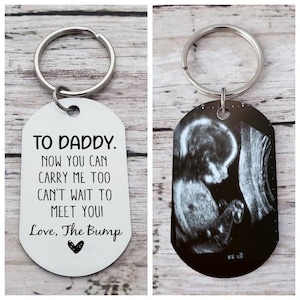 May include: White metal keychain with a silver key ring. The keychain has a black and white ultrasound image and text that reads "To Daddy. Now you can carry me too. Can't wait to meet you! Love, The Bump".