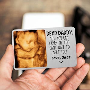 Peut inclure: Une carte de portefeuille en métal argenté avec une image d'échographie et le texte "DEAR DADDY, NOW YOU CAN CARRY ME TOO CANT WAIT TO MEET YOU! Love, Jace". La carte est tenue dans la main d'une personne.