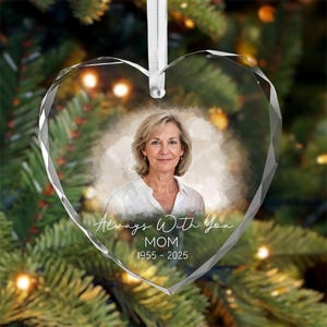 Peut inclure: Un ornement en verre transparent en forme de cœur avec des bords facettés, présentant le portrait d'une femme en chemise blanche. Le texte indique Always With You, MOM, et 1955 - 2025. Un ruban blanc est fixé au sommet pour la suspension.