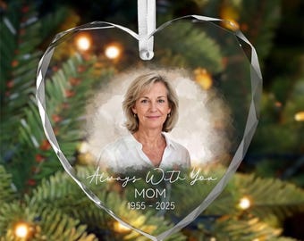 May include: A heart-shaped clear glass ornament with faceted edges, featuring a portrait of a woman in a white shirt. The text reads Always With You, MOM, and 1955 - 2025. A white ribbon is attached to the top for hanging on a tree branch.