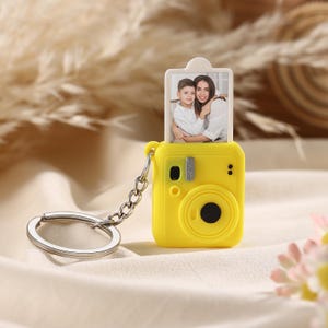 Peut inclure: Un porte-clés jaune en forme d'appareil photo avec une chaîne et un porte-clés argentés. L'appareil photo contient une photo d'une femme et d'un enfant. Le porte-clés est sur une surface blanche avec un arrière-plan flou.