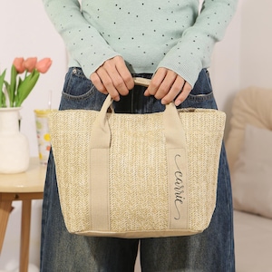 Peut inclure: Un sac fourre-tout beige tiss&eacute;, de forme rectangulaire, avec deux anses. Le sac porte le nom "Carrie" en cursive sur le devant. Le sac est tenu par une personne portant un pull bleu clair et un jean bleu.