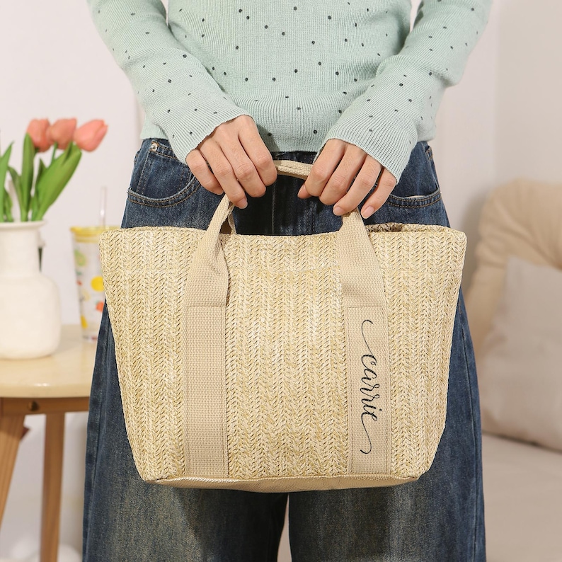 Peut inclure: Un sac fourre-tout beige tiss&eacute;, de forme rectangulaire, avec deux anses. Le sac porte le nom "Carrie" en cursive sur le devant. Le sac est tenu par une personne portant un pull bleu clair et un jean bleu.