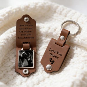 May include: A brown leather keychain with a silver key ring. The keychain has the text "Love You Daddy" and baby footprints. The other part of the keychain has a baby ultrasound photo and the text "Now you can carry me too. Can't wait to meet you! Love, little Bean."