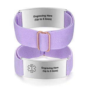 May include: Two lavender medical alert bracelets. Each bracelet has a silver plate with the text "Engraving Here (Up to 5 lines)". One bracelet has a medical symbol. The bands are made of a woven elastic material and are connected by a rose gold-colored buckle.