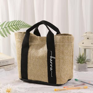Peut inclure: Un sac fourre-tout beige tiss&eacute; avec des poign&eacute;es et des accents noirs. Le sac porte le nom "hanna" en &eacute;criture blanche sur le devant. Le sac est rectangulaire avec un fond plat et est pos&eacute; sur une nappe en dentelle blanche.