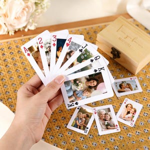 Puede incluir: Una baraja de cartas con fotos personalizadas, sostenida en una mano. Las cartas están abanicadas, mostrando varias fotos familiares. También hay una caja de madera con la inscripción "Love you forever", junto con cartas de fotos adicionales.