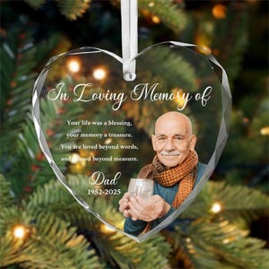 May include: A heart-shaped glass ornament featuring the text In Loving Memory of, a poem, the word Dad, and the years 1952-2025. The ornament displays a photograph of a person wearing a brown scarf and holding a glass, suspended by a white ribbon.