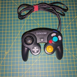 May include: Black Nintendo GameCube controller with a black cable and a red rubber band. The controller has a directional pad, analog stick, and coloured buttons: teal, red, and yellow. The text "NINTENDO GAMECUBE" is printed on the controller.