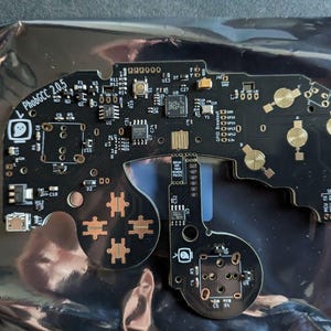 May include: A black circuit board with gold-colored components and the text "PhobGCC 2.0.5". The board has various electronic components, including buttons, chips, and connectors, arranged in a gamepad-like layout.
