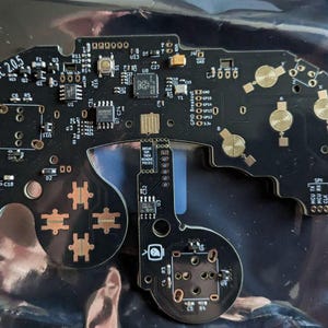 Puede incluir: Placa de circuito negra con componentes de color cobre y el texto "PhobGCC 2.0.5". La placa tiene varios componentes electrónicos, incluyendo chips, botones y conectores. El diseño es intrincado y detallado.