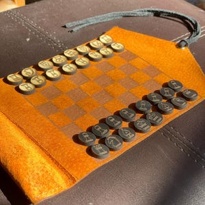 May include: A brown leather travel chess set with black and white pieces. The set is rolled up and has a strap to keep it closed.