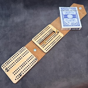 Könnte beinhalten: Ein hölzernes Cribbage-Spielbrett mit einem Kartenspiel in einem braunen Lederetui. Das Cribbage-Spielbrett hat mehrere Reihen von Löchern für Stifte und ein dekoratives Design. Das Etui ist offen und zeigt das Spielbrett und die Karten.