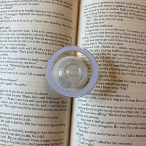 K&ouml;nnte beinhalten: Eine transparente, kreisf&ouml;rmige Lupe mit helllila Rand liegt auf einem offenen Buch. Die Seiten des Buches zeigen gedruckten Text. Die Lupe ist zentriert, mit einer transparenten, kreisf&ouml;rmigen Linse.