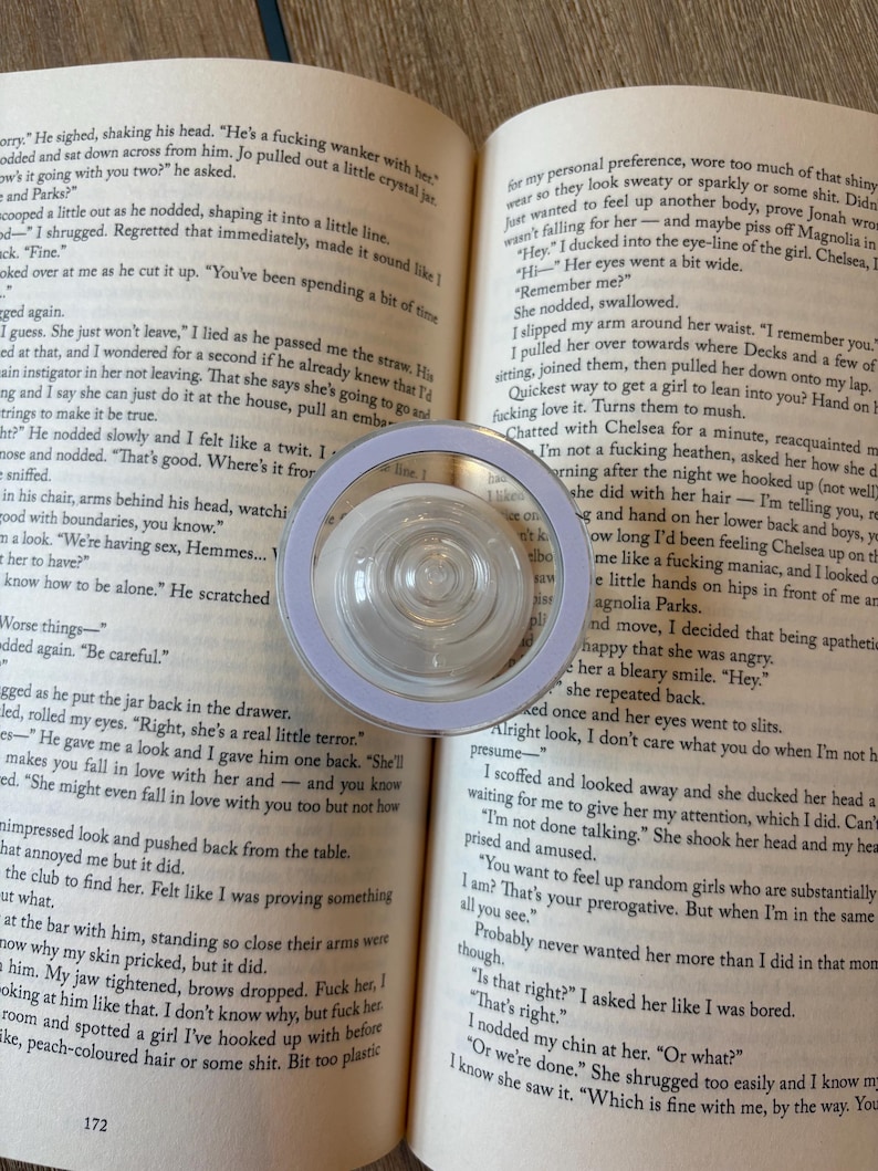 K&ouml;nnte beinhalten: Eine transparente, kreisf&ouml;rmige Lupe mit helllila Rand liegt auf einem offenen Buch. Die Seiten des Buches zeigen gedruckten Text. Die Lupe ist zentriert, mit einer transparenten, kreisf&ouml;rmigen Linse.