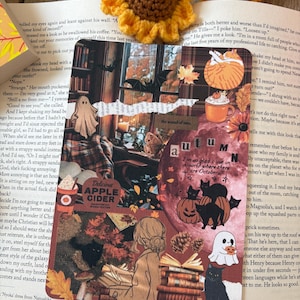 May include: A decorative bookmark with an autumnal theme, featuring illustrations of pumpkins, black cats, ghosts, and fall foliage. The bookmark has the word "autumn" and the text "Delicious Apple Cider".