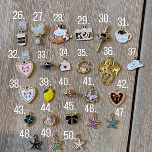 May include: A selection of small, colourful charms featuring diverse designs such as a coffee cup, a cat reading, a camera, a dragon, and a four-leaf clover. The charms are made of metal and enamel, and are displayed on a wooden surface.