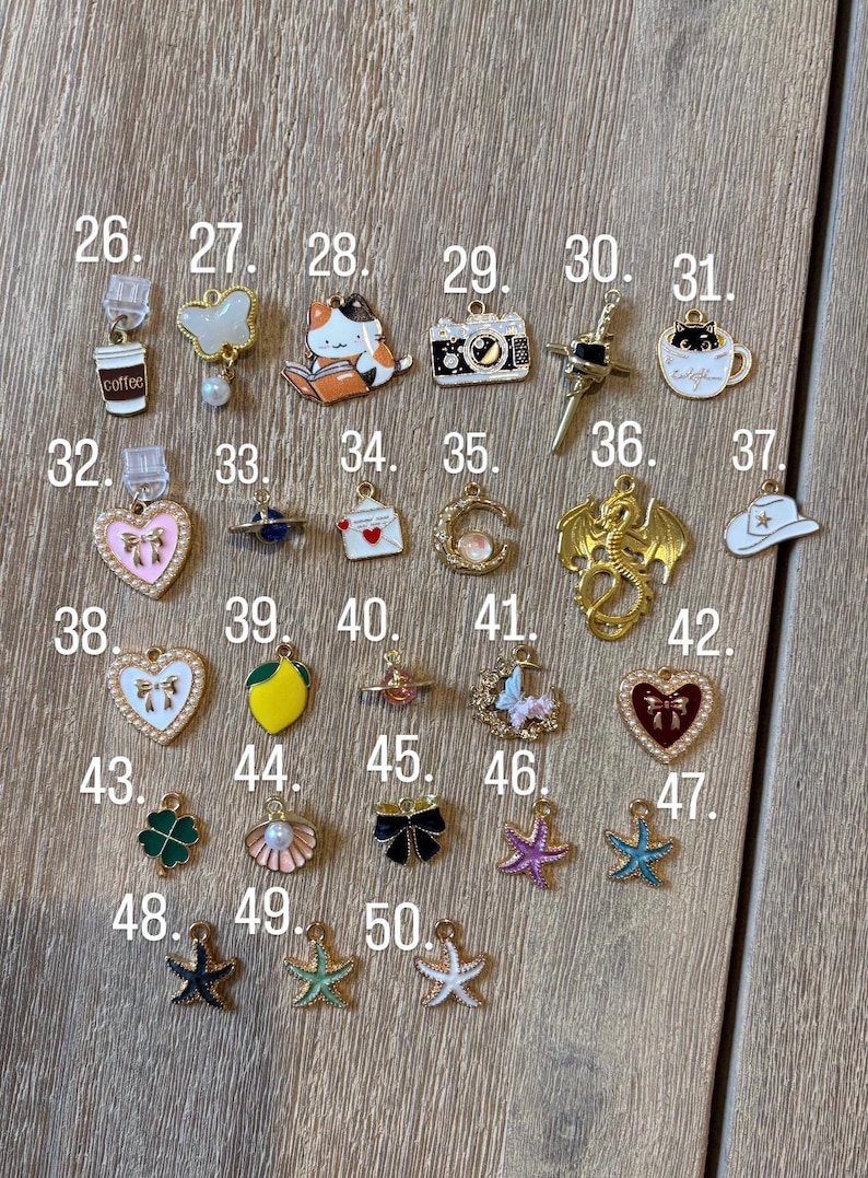 May include: A selection of small, colourful charms featuring diverse designs such as a coffee cup, a cat reading, a camera, a dragon, and a four-leaf clover. The charms are made of metal and enamel, and are displayed on a wooden surface.