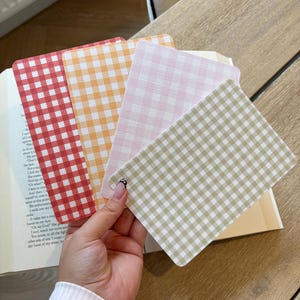 May include: A hand displays four rectangular cards with a gingham pattern in red, orange, pink, and green. The cards are held over an open book. The cards are a variety of colours.