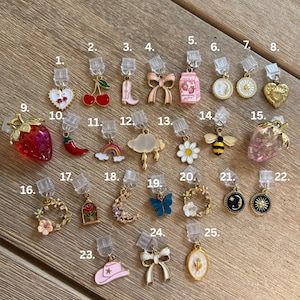 May include: A selection of 25 decorative charms with diverse designs, such as hearts, cherries, bows, a cowboy boot, a milk carton, a coin, a sun, a heart, a strawberry, a chilli pepper, a rainbow, a cloud, a daisy, a bee, a butterfly, a moon, a wreath, and a hat.