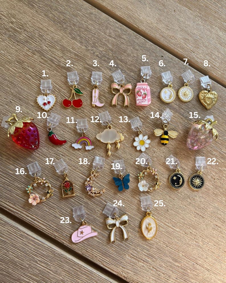 May include: A selection of 25 decorative charms with diverse designs, such as hearts, cherries, bows, a cowboy boot, a milk carton, a coin, a sun, a heart, a strawberry, a chilli pepper, a rainbow, a cloud, a daisy, a bee, a butterfly, a moon, a wreath, and a hat.