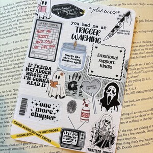 May include: A selection of book-themed stickers, including ghost illustrations, phrases like "Trigger Warning" and "Emotional Support Kindle," and crime scene tape. The stickers are displayed on an open book.