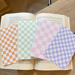 May include: Four cards with a checkerboard pattern in orange, green, pink, and blue are placed on an open book. The cards feature a classic checkerboard design. The book's pages are yellowed.