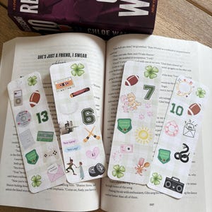 May include: Four decorative bookmarks with various designs, including a cassette tape, football, and the numbers 6, 7, 10, and 13. The bookmarks are placed on an open book with the title "She's Just a Friend, I Swear". A book with the title "Releasing" is in the background.