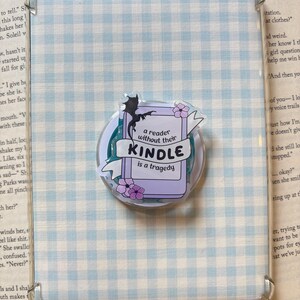 May include: A clear protective case for an e-reader featuring a light blue and white gingham pattern. Attached to the centre is a circular accessory with a graphic of a purple book, a black dragon silhouette, and pink flowers with the text: a reader without their KINDLE is a tragedy.