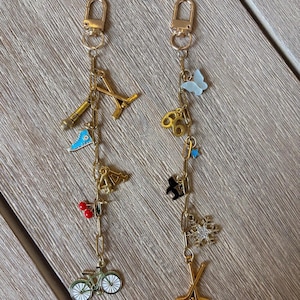 May include: Two gold-toned metal keychains labelled 1 and 2, each featuring a lobster clasp and a vertical chain adorned with various charms. Keychain 1 includes a microphone, blue sneaker, number 44, red cherries, and a bicycle. Keychain 2 features a light blue butterfly, number 66, a small blue star, a black cat, a snowflake, and crossed hockey sticks.
