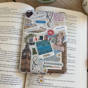 May include: A clear phone case decorated with stickers, including a Big Ben image, a stethoscope, and the words "HATES" and "Baby Ha&iuml;tes." A daisy charm hangs from the case. The case is placed on an open book.