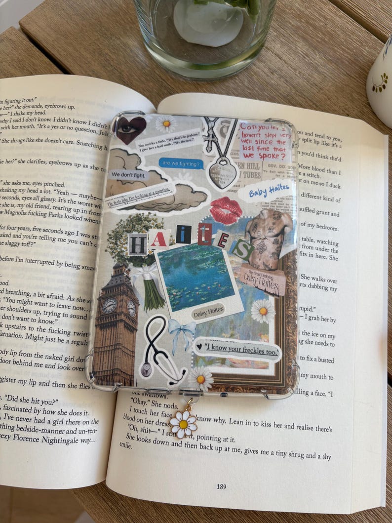 May include: A clear phone case decorated with stickers, including a Big Ben image, a stethoscope, and the words "HATES" and "Baby Ha&iuml;tes." A daisy charm hangs from the case. The case is placed on an open book.