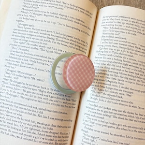 K&ouml;nnte beinhalten: Offenes Buch mit Text und ein kleiner, runder Beh&auml;lter mit rosa-wei&szlig;em Karomuster. Ein hellgr&uuml;ner Beh&auml;lter ist ebenfalls sichtbar. Das Buch liegt auf einer dunklen Holzoberfl&auml;che.