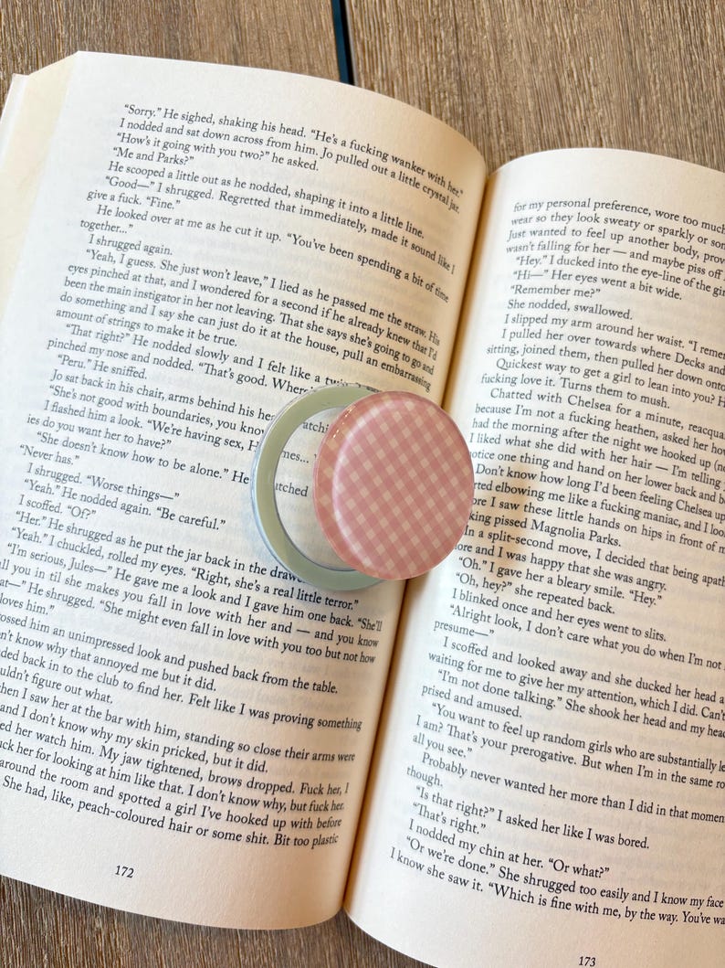 K&ouml;nnte beinhalten: Offenes Buch mit Text und ein kleiner, runder Beh&auml;lter mit rosa-wei&szlig;em Karomuster. Ein hellgr&uuml;ner Beh&auml;lter ist ebenfalls sichtbar. Das Buch liegt auf einer dunklen Holzoberfl&auml;che.