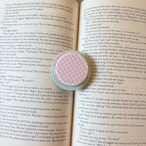 K&ouml;nnte beinhalten: Ein runder Kompaktspiegel mit rosa-wei&szlig;em Vichy-Muster und mintgr&uuml;nem Rand liegt auf einem offenen Buch. Die Seiten des Buches sind vergilbt, und Text ist sichtbar.