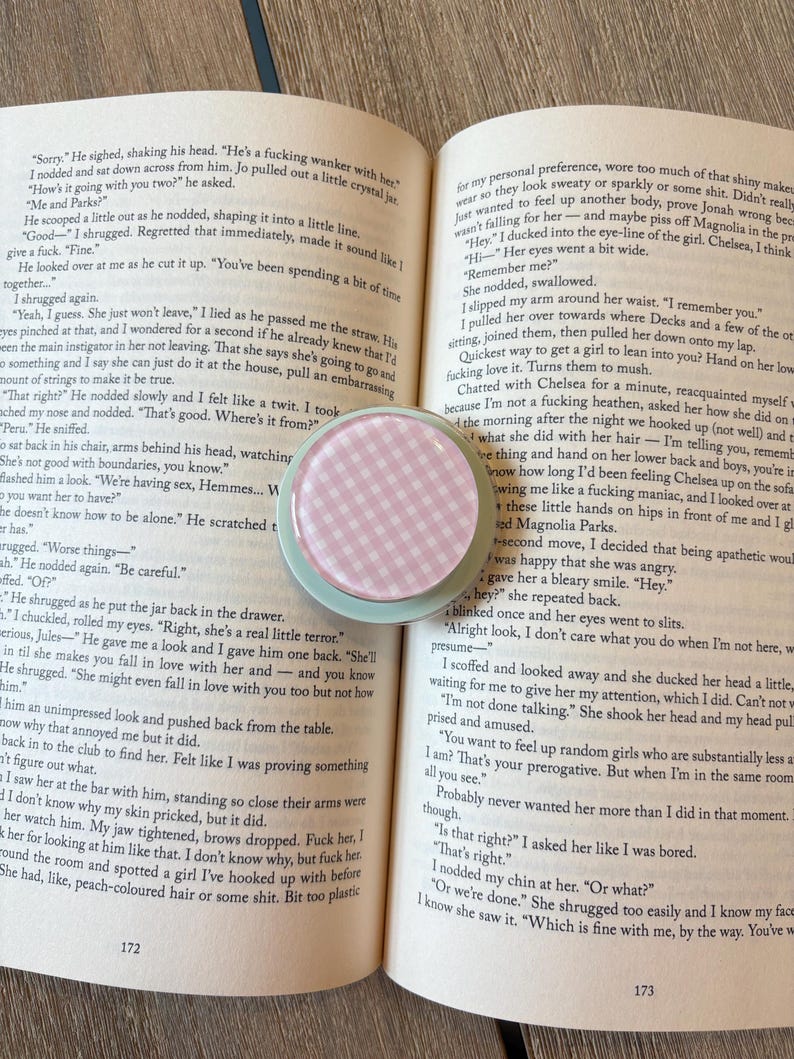 K&ouml;nnte beinhalten: Ein runder Kompaktspiegel mit rosa-wei&szlig;em Vichy-Muster und mintgr&uuml;nem Rand liegt auf einem offenen Buch. Die Seiten des Buches sind vergilbt, und Text ist sichtbar.