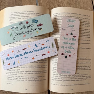 May include: Three rectangular paper bookmarks with printed designs. One is light green with the text Twilight Reading club, one is light blue with a plaid pattern and the text Hoa Hoa Hoa Season, and one is a pink library card with the text This is the bookmark of a killer, Bella.