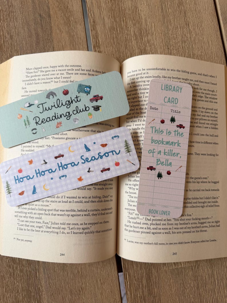 May include: Three rectangular paper bookmarks with printed designs. One is light green with the text Twilight Reading club, one is light blue with a plaid pattern and the text Hoa Hoa Hoa Season, and one is a pink library card with the text This is the bookmark of a killer, Bella.