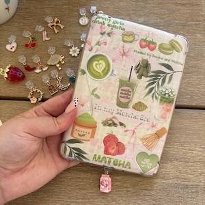 May include: A clear phone case with a matcha theme, featuring text that reads "pretty girls drink matcha" and "In my Matcha Era." The case is decorated with matcha drinks, strawberries, and floral designs. A variety of charms are scattered around the case.
