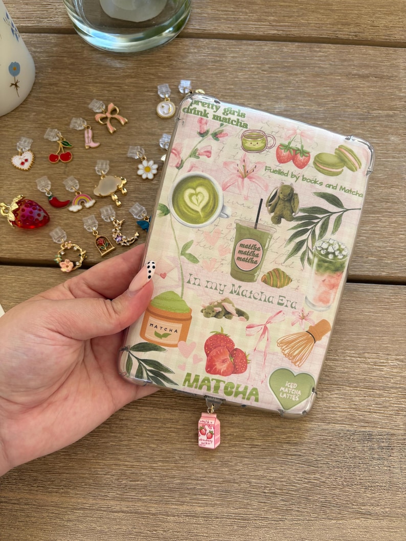 May include: A clear phone case with a matcha theme, featuring text that reads "pretty girls drink matcha" and "In my Matcha Era." The case is decorated with matcha drinks, strawberries, and floral designs. A variety of charms are scattered around the case.