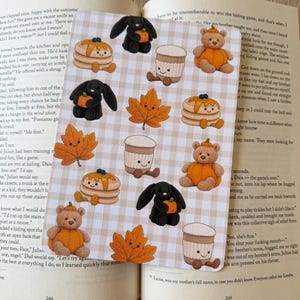 May include: A rectangular sticker sheet with a light beige and white chequered pattern featuring repeating illustrations of black bunnies, brown teddy bears, orange maple leaves, stacks of pancakes, and coffee cups, all with smiling faces.