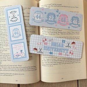 May include: A collection of hockey-themed bookmarks in light blue and white. One bookmark shows a jersey with the number 44, another has "Briar Hockey." Other bookmarks feature jerseys with numbers 22, 66, and 46. The bookmarks are on a book.