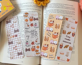 May include: Four decorative bookmarks with fall-themed designs, including ghosts, pumpkins, teddy bears, and the text "Haunted by my TBR" and "Cozy Season". The bookmarks are placed on an open book with crocheted sunflowers.